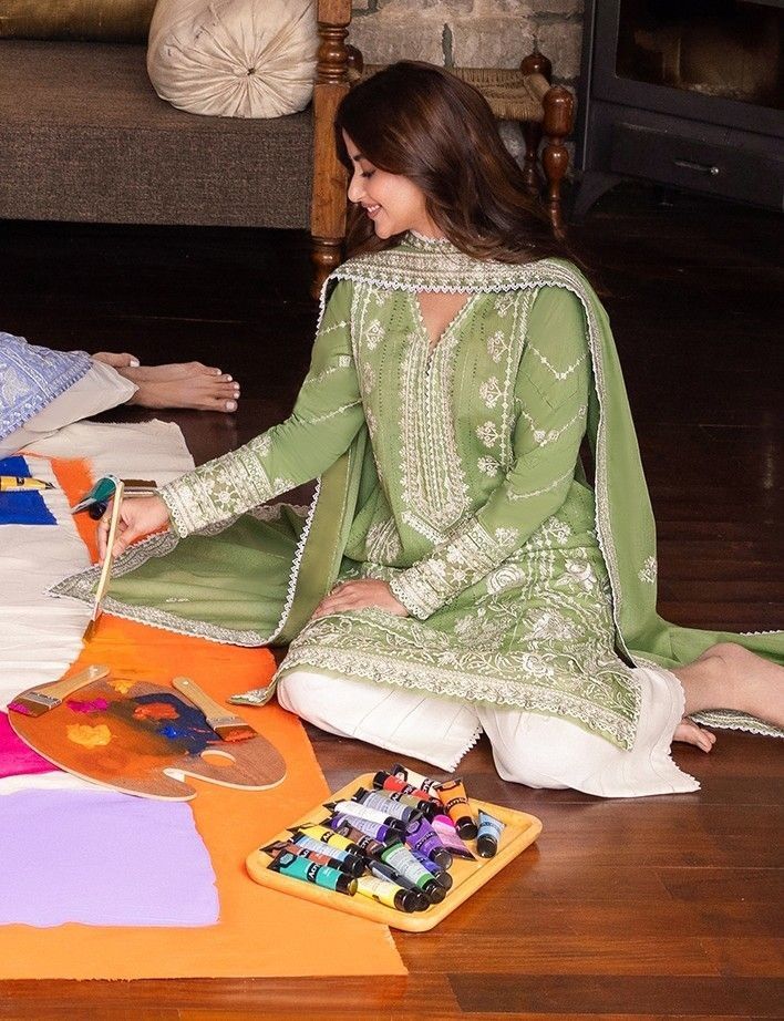 A woman in a light green embroidered traditional outfit sits on a wooden floor, painting with a paintbrush. She is surrounded by art supplies, including a paint palette and a tray of colorful paint tubes, in a cozy indoor setting.