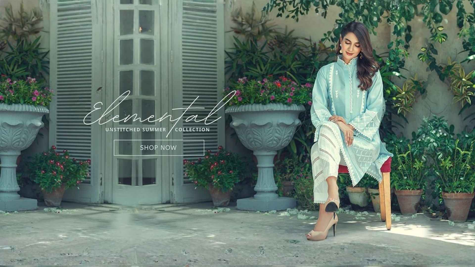 A woman sits on a wooden chair in an outdoor courtyard setting, wearing a light blue and white embroidered unstitched summer collection outfit by Elemental. The scene features large stone planters with pink flowers and a rustic gray door in the background.